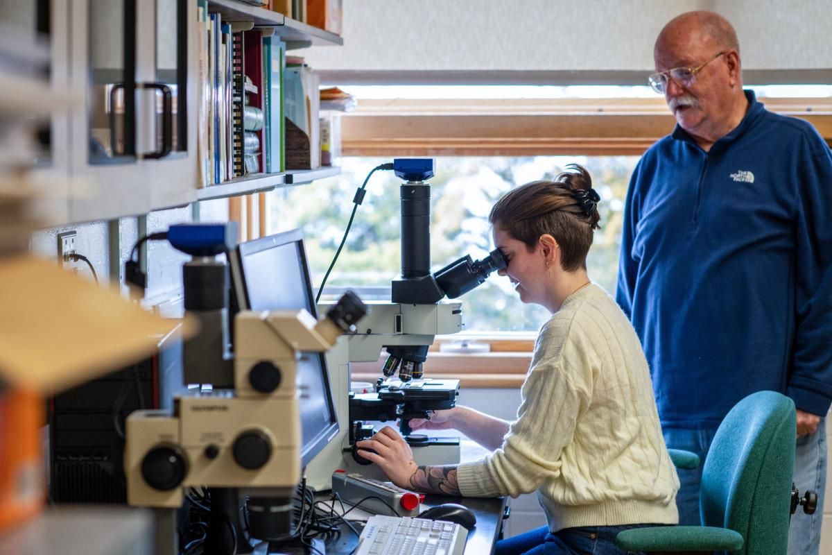 Emma Gallagher seated looking into a microscope with guidance from Dr. Jeff Turner