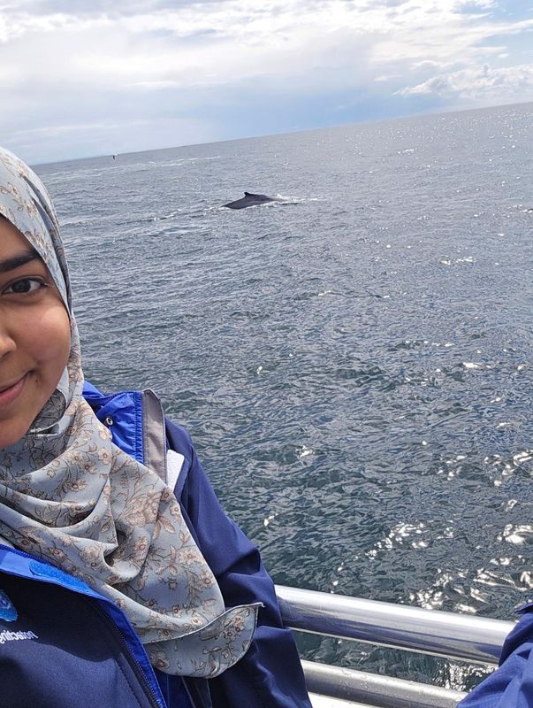 Eman Khwaja standing on a boat with a humpback whale in the ocean behind her.