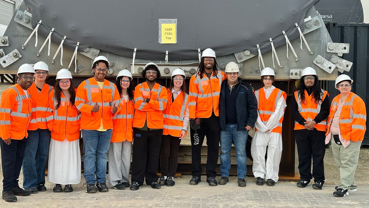 UMass Dartmouth and New Bedford High School students wearing hard hats and reflective vests and standing in front of a wind turbine element.