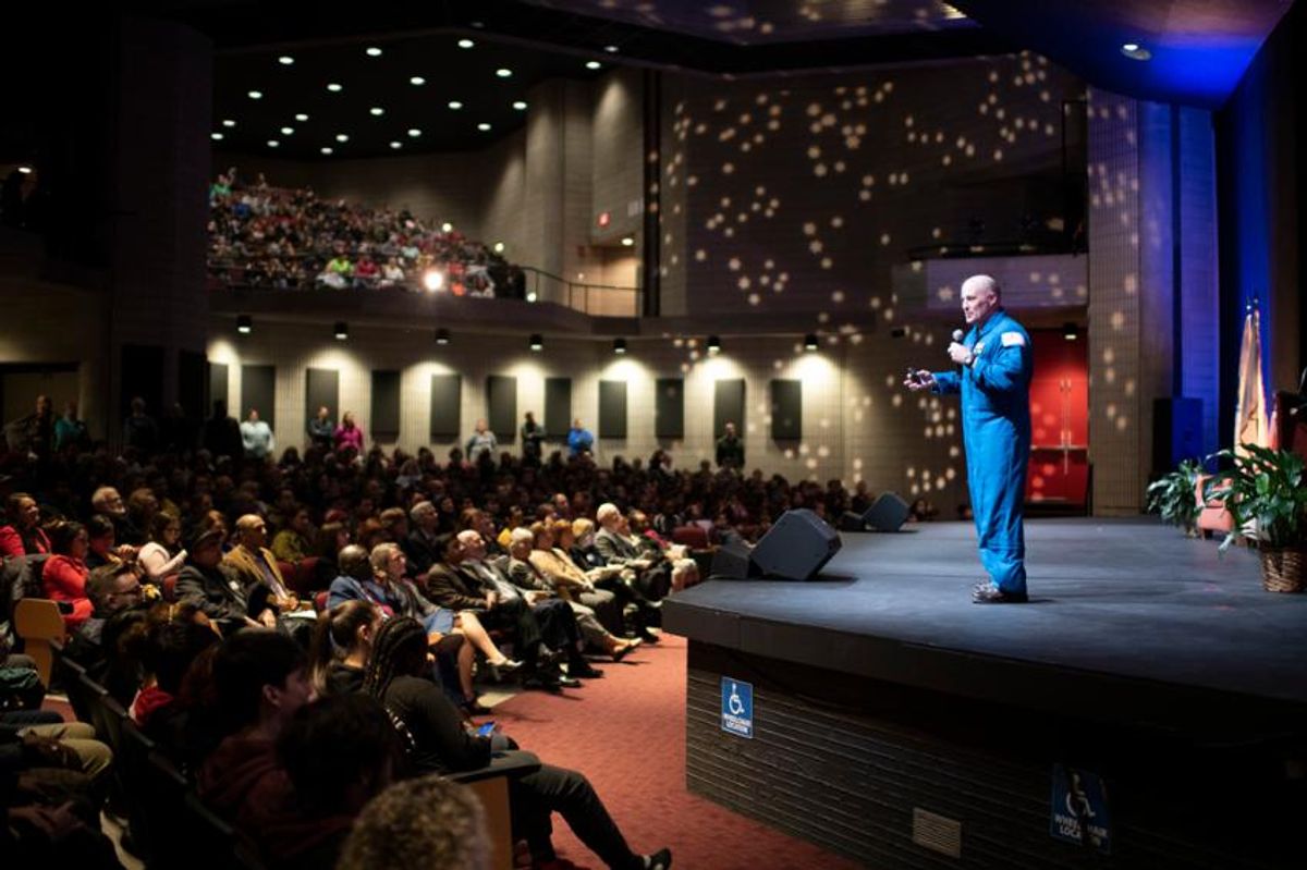 Scott Tingle speaking on stage in his NASA flight suit with stars projected on walls
