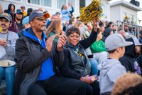 A cheerful couple sits close together in the stands at the Blue & Gold Weekend football game, Behind them, a crowd of spectators fills the bleachers.