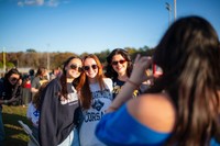 Three individuals wearing UMass Dartmouth Corsairs gear smile and pose for a photo during the Blue & Gold Family Festival, while another person takes their picture