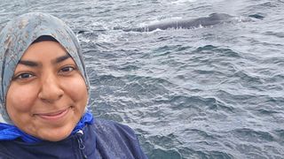 Eman Khwaja standing on a boat with a whale surfacing in the background