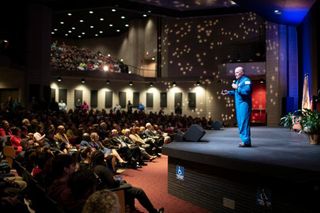 Scott Tingle speaking on stage in his NASA flight suit with stars projected on walls
