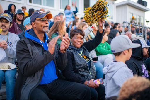 A cheerful couple sits close together in the stands at the Blue & Gold Weekend football game, Behind them, a crowd of spectators fills the bleachers.