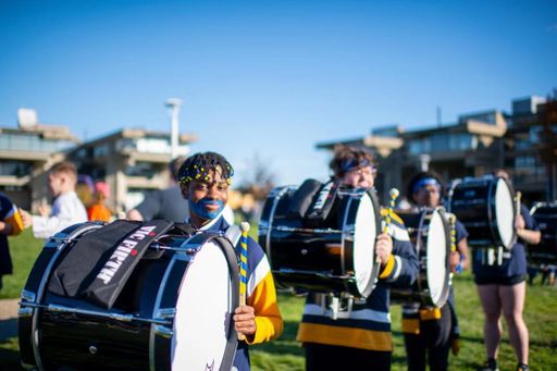 A student drummer with blue face paint smiles while holding drumsticks in front of a bass drum during the Blue & Gold Family Festival