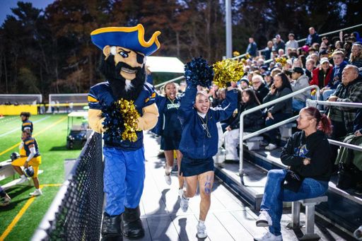 Arnie walks along the bleachers at the Blue & Gold Weekend football game, holding pom-poms and accompanied by excited cheerleaders as the crowd watches from the stands