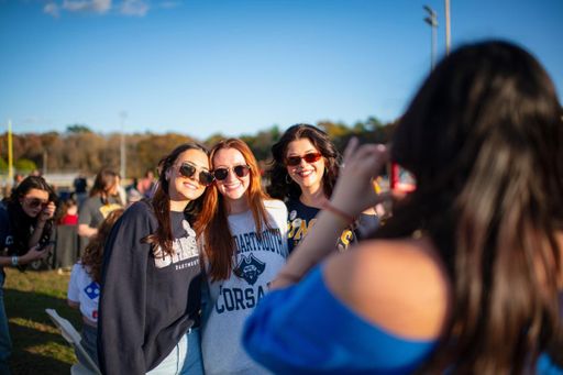 Three individuals wearing UMass Dartmouth Corsairs gear smile and pose for a photo during the Blue & Gold Family Festival, while another person takes their picture