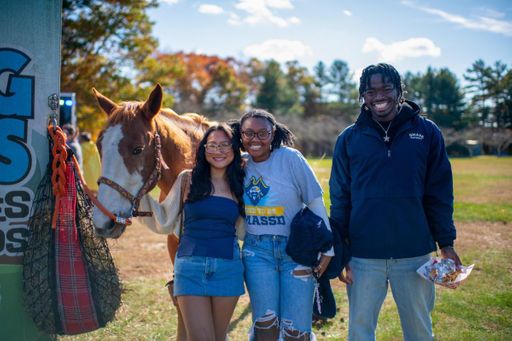 Three students pose next to a horse at the Blue & Gold Family Festival, one wears a UMass Dartmouth t-shirt reading Proud to be UMassD