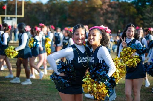 Two Corsair cheerleaders with gold and blue pom-poms pose and smile at the football game with a group of cheerleaders in the background wearing matching uniforms and pink bows