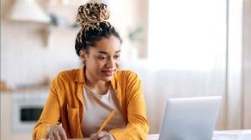 Stylish african-american female student with afro dreadlocks, studying while using a laptop, taking notes on notepad and smiling..
