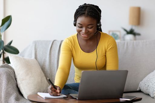 Girl wearing headphones sitting on the couch taking notes while on the computer.