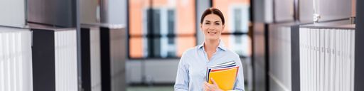 brunette teacher holding folders and notebooks while standing in school corridor