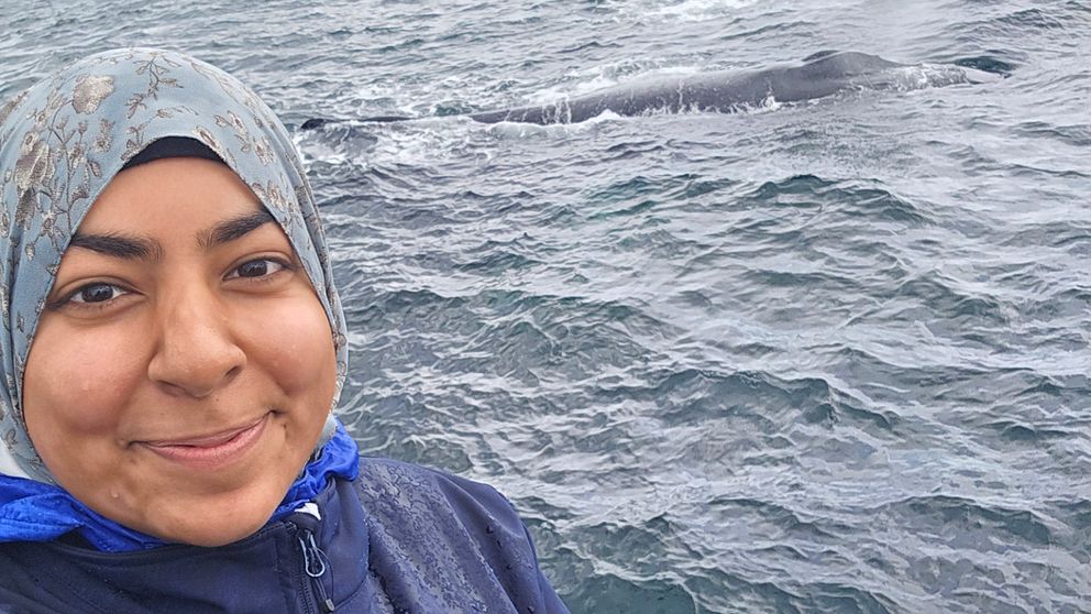 Eman Khwaja standing on a boat with a whale surfacing in the background